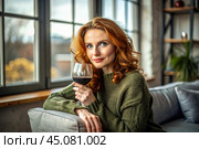 Close-up. A modern, stylish, beautiful woman with red hair and a glass of red wine in an armchair in the living room of the house. Стоковое AI-изображение, AI-художник Photojuli / Фотобанк Лори