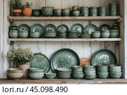 A wooden shelf with a set of green ceramic dishes with a craquelure effect (cracks on the glaze surface). The shelf is made in a vintage style, with noticeable signs of wear and tear, which gives the whole composition a rustic and country character. In the center of the composition are bowls, plates and cups of various sizes, neatly stacked. To the right and left are ceramic pots with plants and flowers, complementing the atmosphere of coziness and natural simplicity. Стоковое AI-изображение, AI-художник Анна Кривицкая / Фотобанк Лори