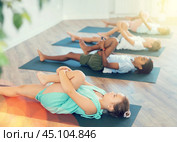 Children doing yoga with group in studio, lying on mat with knee pressed to chest with hands, performing hamstring and low back stretching Ardha Pawanmuktasana. Стоковое фото, фотограф Яков Филимонов / Фотобанк Лори
