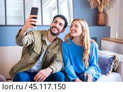 Happy young couple takes a selfie together with smartphone on sofa at home. Стоковое фото, фотограф Raquel Corcoles Mendez / Фотобанк Лори