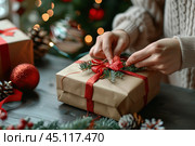 Woman in sweater is packing a gift box. Female hands tying red ribbon, bow around giftbox wrapped into craft paper, preparing Xmas presents. Christmas tree festive background. Close up. New year DIY. Стоковое AI-изображение, AI-художник Марина Демидюк / Фотобанк Лори