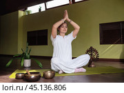 Woman practicing meditation in Kundalini yoga with welcoming namaste gesture and hands raised up, sitting in comfortable lotus position on mat in room, wearing white clothes. Стоковое фото, фотограф Сергей Чайко / Фотобанк Лори