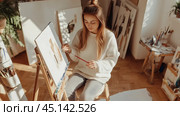 Young woman painting on an easel in a sunlit art studio filled with canvases, brushes, and creative materials during the afternoon. Стоковое AI-изображение, AI-художник Сергей Толмачёв / Фотобанк Лори