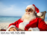 A portrait of satisfied, cheerful Santa Claus in dark glasses resting in beach lounger on a tropical beach. Стоковое AI-изображение, AI-художник Михаил Строгалев / Фотобанк Лори
