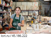 Young female worker in pottery factory lowers fired clay plates and cups into container with glaze. Стоковое фото, фотограф Яков Филимонов / Фотобанк Лори