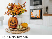 A vase of flowers,a jack pumpkin and candles on a tray. In the background - the interior of a white kitchen in Scandi style. The concept of home and comfort. Autumn decor for the Halloween holiday. Стоковое фото, фотограф Photojuli / Фотобанк Лори