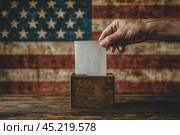 Close-up human hand with vote bulletin and election box on the background of USA flag. Ballot paper for Democrats and Republicans. Elections, election campaign. Стоковое AI-изображение, AI-художник Ирина Михайличенко / Фотобанк Лори