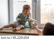 A cute 2-year-old boy in a cap is playing on the windowsill with a wooden airplane. Children's wooden toys for child development. Стоковое фото, фотограф Photojuli / Фотобанк Лори