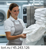A woman working in a laundry or hotel industry. She is wearing a uniform with an apron and is carefully folding white bed linen. Industrial washing machines are visible in the background, which emphasizes the professional environment. The woman is smiling, creating a sense of goodwill and neatness in her work. Стоковое AI-изображение, AI-художник Анна Кривицкая / Фотобанк Лори