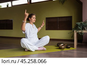 Woman kundalini yoga, doing Adi Shakti meditation, sitting on a mat in a lotus position with her hands raised up and a gesture with her index finger raised up, practicing in white clothes in a room. Стоковое фото, фотограф Сергей Чайко / Фотобанк Лори