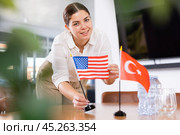 Positive young woman putting flag of the USA on the table with flag of Turkey in conference room. Стоковое фото, фотограф Яков Филимонов / Фотобанк Лори