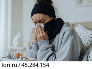 Young sick woman with cold or flue sitting on grey couch, holding a tissue to clear, blowing runny nose. Sneezing Girl under gray plaid blanket with handkerchief to dripping nose, blowing snot at home. Стоковое AI-изображение, AI-художник Марина Демидюк / Фотобанк Лори