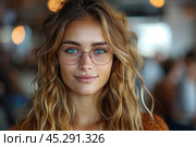 Portrait of a beautiful young woman with glasses, wavy hair and blue eyes in casual clothes smiling at the camera while standing against blurred background of modern cafe interior. Cope space. Banner. Стоковое AI-изображение, AI-художник Евгений Демидюк / Фотобанк Лори