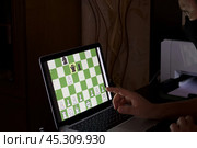 A man in a medical mask solves a chess problem online. Chess board with figures on a laptop screen. Leisure in self-isolation, in the twilight. International chess day. Стоковое фото, фотограф Vadzim Yakubovich / Фотобанк Лори