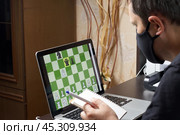 A man in a medical mask solves a chess problem online. Chess board with figures on a laptop screen. Leisure in self-isolation, in the twilight. International chess day. Стоковое фото, фотограф Vadzim Yakubovich / Фотобанк Лори