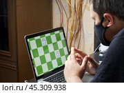 A man in a medical mask solves a chess problem online. Chess board with figures on a laptop screen. Leisure in self-isolation. International chess day. Стоковое фото, фотограф Vadzim Yakubovich / Фотобанк Лори