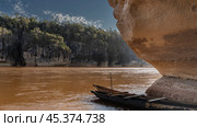 Canoes hollowed out of wood are moored at the entrance to the cave. The rock overhangs the red-brown river. Green vegetation on the far steep bank. Blue sky, clouds. Madagascar. Manambolo River. Стоковое фото, фотограф Вера Ивановна Кубарева / Фотобанк Лори