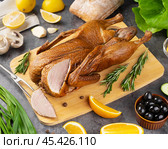 Deliciously roasted duck served on a wooden board with fresh herbs and citrus slices in a well-lit kitchen setting. Стоковое фото, фотограф Евгения Трушкова / Фотобанк Лори