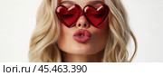 A delicate blonde with a beautiful, refined appearance. Her gaze is full of light and a touch of mystery. Vivid crimson lips complete the look, drawing attention. Red heart-shaped glasses add a playful and romantic touch. Стоковое AI-изображение, AI-художник Ярославский Максим / Фотобанк Лори