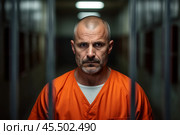 A prisoner in an aged orange uniform looks directly at the camera from behind metal bars in a dimly lit detention center, capturing the gravity of incarceration. Стоковое AI-изображение, AI-художник Sergey Grishin / Фотобанк Лори