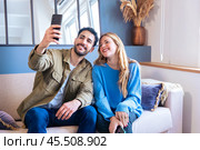 Happy young couple takes a selfie together with smartphone on sofa at home. Стоковое фото, фотограф Raquel Corcoles Mendez / Фотобанк Лори