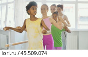 Multiethnic group of teenage girls and boys, ballet class students, practicing demi plie at barre during lesson for beginners under guidance of female teacher in light-filled choreography studio. Стоковое видео, видеограф Яков Филимонов / Фотобанк Лори