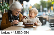 Grandmother enjoys cake with small child at cafe in the afternoon sunlight during a joyful family outing. Стоковое AI-изображение, AI-художник Сергей Толмачёв / Фотобанк Лори