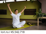 Positive woman doing ego or personality mudra, healthy lifestyle and practicing Kundalini yoga sitting in lotus position with closed eyes on mat in meditation room. Стоковое фото, фотограф Сергей Чайко / Фотобанк Лори