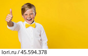 A happy boy wearing a white shirt and a yellow bowtie on a yellow background, gestures a thumbs-up gesture. Copy space. Стоковое AI-изображение, AI-художник Михаил Строгалев / Фотобанк Лори