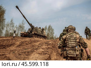САУ МСТА-С стоит на возвышенности, по которой поднимаются солдаты вооруженных сил армии России. Гаубица и пехота на позиции. Штурмовой отряд проходит мимо артиллерийского расчёта. (2019 год). Редакционное фото, фотограф Евгений Зимин / Фотобанк Лори