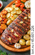 Grilled flank steak with roasted potatoes, garlic, and cherry tomatoes on wooden platter. Стоковое AI-изображение, AI-художник Юрий Поздников / Фотобанк Лори