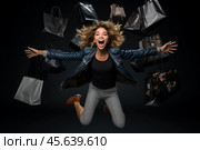 A woman with curly hair joyfully jumps in the air, surrounded by various shopping bags, expressing excitement and happiness in a darkened environment. Стоковое AI-изображение, AI-художник Sergey Grishin / Фотобанк Лори