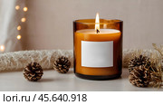 A mock-up of a cozy burning candle made of amber glass with an empty white label in a festive, New Year's home interior. Стоковое AI-изображение, AI-художник Photojuli / Фотобанк Лори