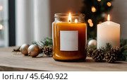 A mock-up of a cozy burning candle made of amber glass with an empty white label in a festive, New Year's home interior. Стоковое AI-изображение, AI-художник Photojuli / Фотобанк Лори
