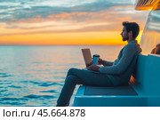 A young guy on a yacht is working on a laptop with a mug of fragrant coffee. Beautiful sea dawn. Стоковое AI-изображение, AI-художник Ирина Ручушкина / Фотобанк Лори