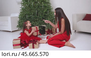 Boxing Day. Mom in red suit and little daughter in red dress open New Year gift box near the Christmas tree and rejoices holidays. Parent with love and tenderness look at kid. Merry Christmas footage. Стоковое видео, видеограф Ирина Михайличенко / Фотобанк Лори