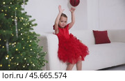 Little girl in red dress sitting on the sofa near the Christmas tree, clapping her hands and rejoicing in Christmas. Footage video. Стоковое видео, видеограф Ирина Михайличенко / Фотобанк Лори