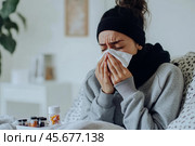 Young sick woman with cold or flue sitting on grey couch, holding a tissue to clear, blowing runny nose. Sneezing Girl under gray plaid blanket with handkerchief to dripping nose, blowing snot at home. Стоковое AI-изображение, AI-художник Марина Демидюк / Фотобанк Лори