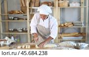 Skilled mature female baker in white uniform carefully slicing dough on flour-covered worktable in cozy bakery filled with freshly baked bread and baking tools. Стоковое видео, видеограф Яков Филимонов / Фотобанк Лори