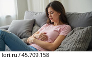 Woman sitting on couch holding stomach in pain, feeling unwell, experiencing abdominal discomfort, health issue, upset stomach, digestive problems, physical discomfort, indoor setting. Стоковое AI-изображение, AI-художник Павел Муравьев / Фотобанк Лори