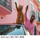 Кот с мужчиной фотографируются на улице. Рыжий кот сидит на крыше автомобиля, подняв лапу, как приветствуем кого-то. Рядом стоит мужчина в розовой рубашке и солнечных очках, делающий селфи с котом на фоне ярко-розовых фасадов зданий. Стоковое AI-изображение, AI-художник Анна Чижова / Фотобанк Лори