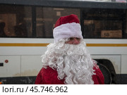 Мужчина в костюме Деда Мороза с посохом идет по улице на фоне рейсового автобуса в городе Московской области, Россия. Редакционное фото, фотограф Николай Винокуров / Фотобанк Лори