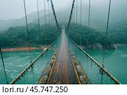 Suspension bridge spanning misty turquoise river amidst lush green mountains. Стоковое AI-изображение, AI-художник Иван Рябоконь / Фотобанк Лори