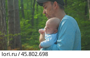 A bearded man in a black baseball cap and blue jersey, carrying a baby in a blue onesie in his arms. He is walking through a green forest. Father and son walking in a park. Side view. Close-up. Стоковое видео, видеограф Воловодов Владимир / Фотобанк Лори