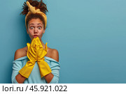 Surprised woman with curly hair and yellow cleaning gloves on blue background. Стоковое AI-изображение, AI-художник Zakharova Natasha / Фотобанк Лори