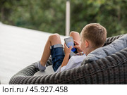 A 5-year-old little boy with a smartphone in his hands on the veranda. The child is playing on the phone. Harm of gadgets for children. Protect your child from the smartphone. Kids and gadgets. Стоковое фото, фотограф Photojuli / Фотобанк Лори