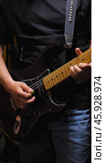 Musician in black clothes and blue jeans playing a black electric guitar with maple deck, green plectrum visible, studded guitar strap attached. Стоковое фото, фотограф Воловодов Владимир / Фотобанк Лори