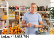 Elderly man with paper list in hands, walks around minimarket, puts groceries in market basket. Стоковое фото, фотограф Яков Филимонов / Фотобанк Лори