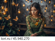 Portrait of a beautiful girl in a green dress among many suspended light bulbs and butterflies fluttering around her. Daydreaming. A magical atmosphere. Стоковое AI-изображение, AI-художник Photojuli / Фотобанк Лори