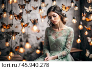 Portrait of a beautiful girl in a green dress among many suspended light bulbs and butterflies fluttering around her. Daydreaming. A magical atmosphere. Стоковое AI-изображение, AI-художник Photojuli / Фотобанк Лори